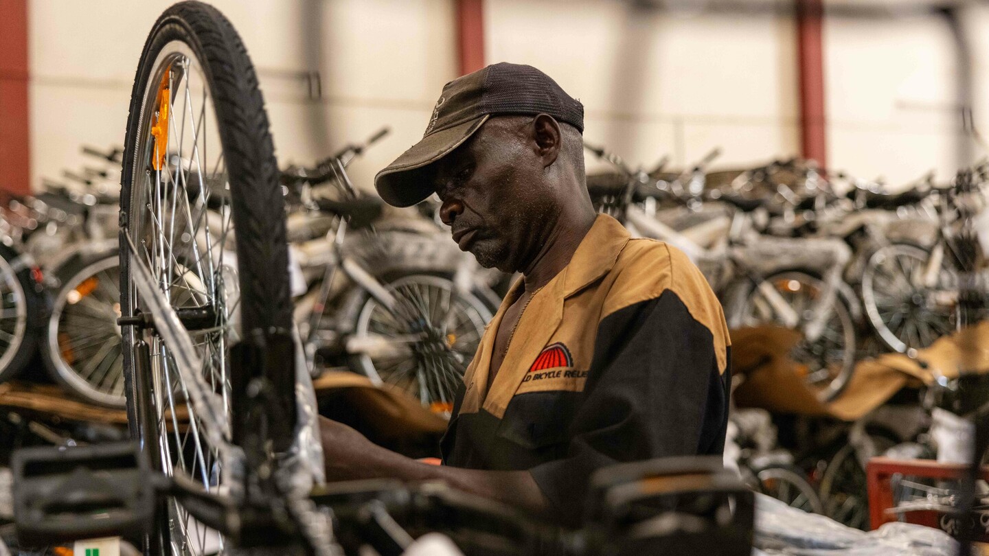 A man in a workshop assembles a bicycle while wearing a cap. Numerous other bicycles are visible in the densely packed background.