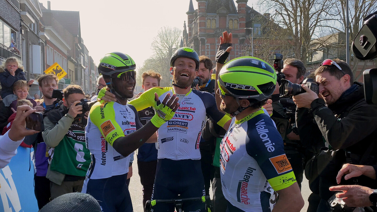 Road cyclists celebrating at the finish line surrounded by photographers and fans.